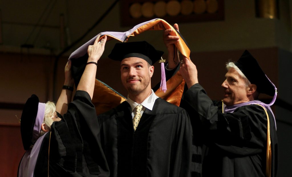 A School of Dentistry Doctor of Dental Surgery student wearing doctoral regalia is hooded by a School of Dentistry faculty member and his father during the 2024 commencement ceremony. 